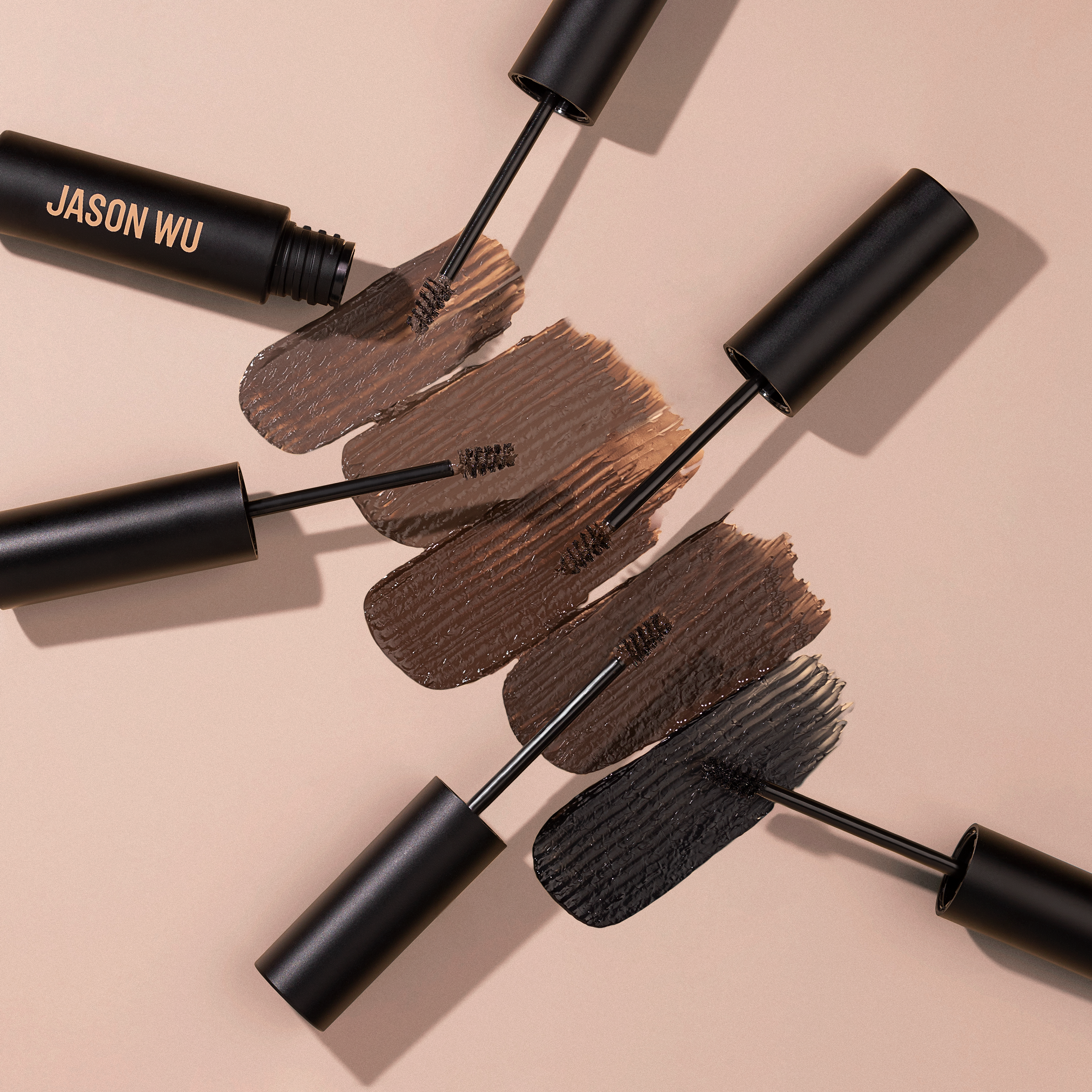 Four open mascara tubes with different shades and brush strokes on a beige background