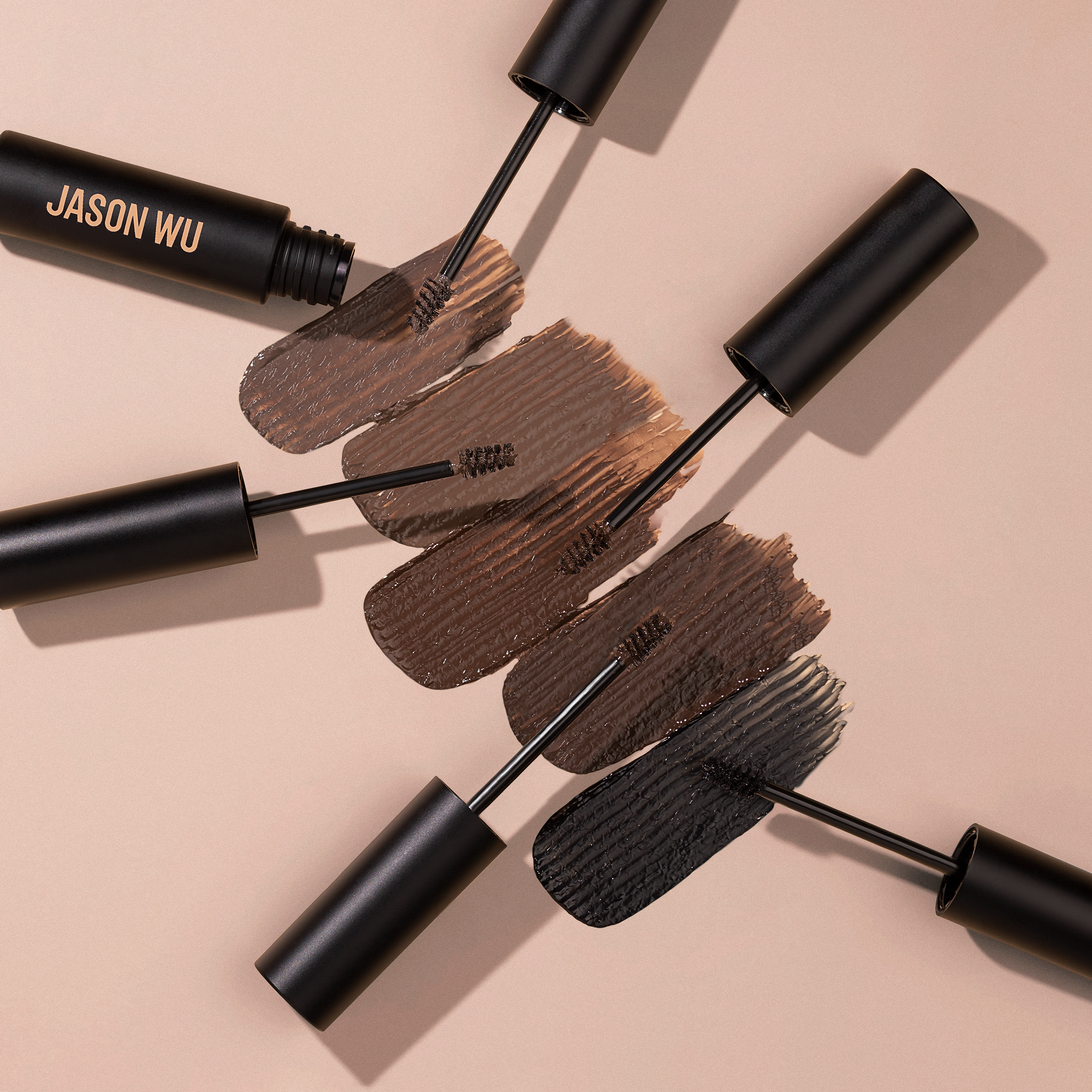 Four makeup brushes with various shades of brown and black brow gel on a beige background.