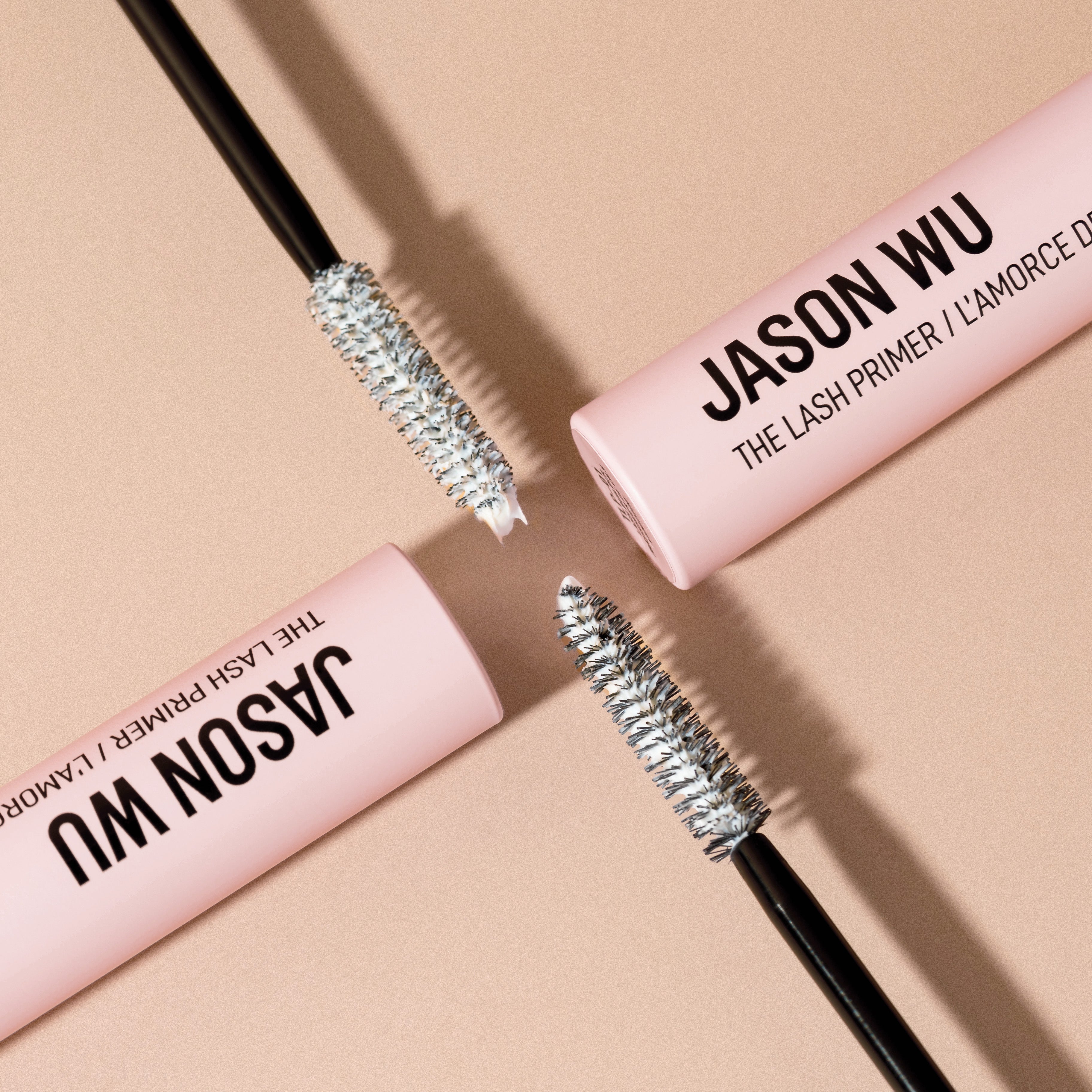 Two mascara wands with silver bristles next to pink tubes labeled 'Jason Wu The Lash Primer' on a beige background.